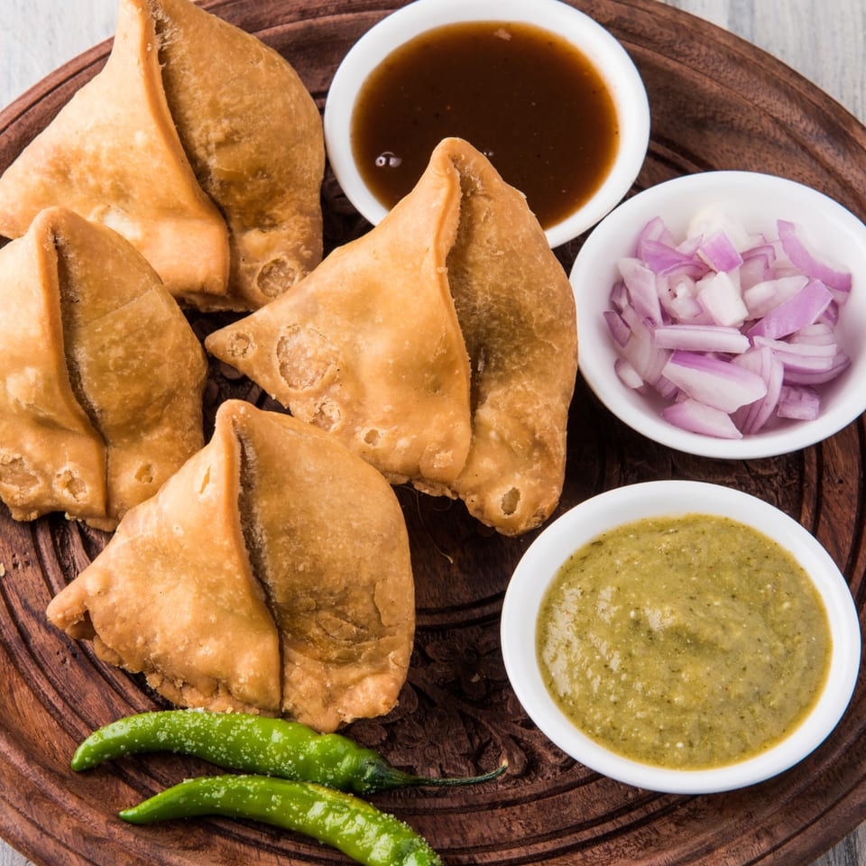 Golden-fried samosas with green chili peppers, brown chutney, green chutney, and chopped red onions.